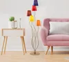 A multi-colored floor lamp stands next to a pink sofa and a small wooden side table with a cactus and a vase on it. Produktbild-Vorschau