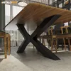 A close-up view of a wooden dining table with black metal legs, set in a dining room with chairs and curtains in the background. Produktbild-Vorschau