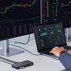 A person's hands are using a laptop connected to a docking station, with multiple monitors displaying financial charts in the background. Produktbild-Vorschau