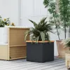A dark gray cube-shaped planter with a wooden top, containing a palm plant, sits on a light gray wooden deck. A wicker sofa with white cushions is visible to the left, and tall bamboo plants in terracotta pots are to the right. Produktbild-Vorschau
