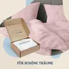 An open cardboard box with bedding inside, placed on a bed with pink bedding. Produktbild-Vorschau