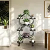 A black metal plant stand with multiple tiers and heart-shaped shelves, filled with various potted plants, placed indoors next to a white cabinet and a wooden table. Náhled obrázku produktu
