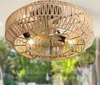 A close-up view of a ceiling fan with a woven rope shade, illuminated by light bulbs, against a blurred background of greenery. Produktbild-Vorschau