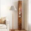 A tall, narrow cabinet with a wood-grain finish is shown next to a floor lamp and a sofa in a room. Podgląd zdjęcia produktu
