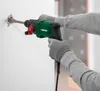 A person wearing gray gloves and a gray sweater is using a green and black rotary hammer drill to break a hole in a white wall. Produktbild-Vorschau