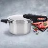A stainless steel pressure cooker is shown on a textured gray surface, with raw meat and spices in the background. Podgląd zdjęcia produktu
