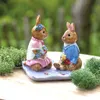Two ceramic bunny figurines sitting on a decorative mat, surrounded by greenery. Náhled obrázku produktu