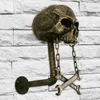 A skull-shaped wall mount with chains and wrenches, attached to a brick wall. Produktbild-Vorschau