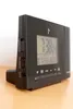 A black digital clock with a projection feature sits on a wooden surface against a white background. Produktbild-Vorschau