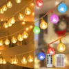 A string of fairy lights with round, textured bulbs in various colors, some warm white and others colored, displayed in a split image showing both indoor and outdoor settings. A remote control and battery pack are also visible. Produktbild-Vorschau