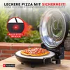 A pizza oven with a pizza on its stone, set outdoors on a wooden table with a brick wall and greenery in the background. Produktbild-Vorschau