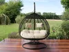 A hanging egg chair with cushions sits on a wooden deck in a garden setting. Náhled obrázku produktu