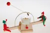 A wooden seesaw with two elf-like figures, a lit candle, and a red ball on a string. Anteprima dell’immagine prodotto