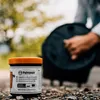 A jar of Petromax care conditioner is placed on the ground in front of a person holding a Dutch oven. Produktbild-Vorschau