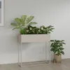 A planter with plants on a stand next to another potted plant, against a light gray wall. Produktbild-Vorschau