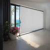A white roller blind is installed on a large glass sliding door overlooking a swimming pool and the ocean. Náhled obrázku produktu