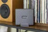 A gray Fujifilm Instax Link Wide printer is placed on a shelf next to a speaker and a stack of vinyl records. Produktbild-Vorschau