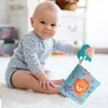A baby sitting on a rug and playing with a soft toy book. Anteprima dell’immagine prodotto