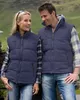 A man and a woman wearing matching navy blue puffer vests and jeans, standing outdoors in a hilly, green landscape. Anteprima dell’immagine prodotto