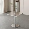 A square metal table with a single pedestal base and a round foot, placed on a tiled floor. A glass vase with a green plant is on the table. Curtains and a decorative pedestal are in the background. Anteprima dell’immagine prodotto