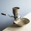 A manual grain mill sits on a wooden surface, with a bowl collecting the ground grain. Produktbild-Vorschau