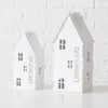 Two white house-shaped lanterns are displayed against a white brick wall. Produktbild-Vorschau