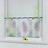 A sheer kitchen curtain with a colorful firework pattern hangs on a rod in a window. A single sunflower rests on the windowsill below. Produktbild-Vorschau
