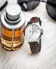 A watch and a bottle of perfume are placed on a white fluffy surface, with sunglasses in the background. Produktbild-Vorschau