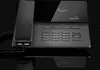 A modern, dark-colored VoIP phone with a handset resting on its base, displayed on a reflective black surface against a black background. Produktbild-Vorschau