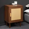 A wooden nightstand with a cane door and a gold handle, placed next to a bed with pillows.