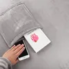 A hand is shown placing a smartphone into a pocket of a gray blanket with sleeves. A book is also visible in the pocket. Anteprima dell’immagine prodotto