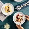 Two bowls of yogurt with fruit and granola are displayed on a dark green tablecloth. One bowl is held by a person's hands, and the other is next to a newspaper and a glass of water. Produktbild-Vorschau
