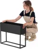 A woman is kneeling next to a black rectangular planter on a stand, with her hands on the planter. Produktbild-Vorschau