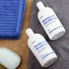 Two bottles of shampoo and conditioner are placed on a gray towel, next to a blue towel and a hairbrush. Podgląd zdjęcia produktu
