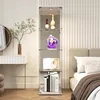 A tall, narrow glass display cabinet with LED lighting, showcasing various items on its shelves, placed in a bedroom setting next to a bed and a vanity. Produktbild-Vorschau