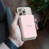A hand holding a pink power bank attached to a gold smartphone, with a blurred background of a chair and a plant. Podgląd zdjęcia produktu