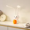 A white rabbit-shaped LED desk lamp with an orange cup holder sits on a white shelf next to a clock and a book. Produktbild-Vorschau