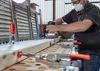 A person is using a sander on a piece of wood that is clamped to a workbench. There are other clamps and tools visible. Náhľad obrázku produktu