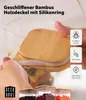 Hand setzt einen Bambusdeckel mit Silikonring auf einen klaren Glas-Lebensmittelaufbewahrungsbehälter, mit zwei weiteren Behältern gefüllt mit Bohnen und Pasta darunter Produktbild-Vorschau