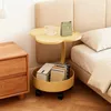 A yellow side table with a flower-shaped top is placed next to a bed and a wooden nightstand. The table has a lower shelf holding books and a box. A mug and a calendar are on the top surface. Produktbild-Vorschau