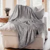 A soft, gray blanket is draped over a white sofa, with a textured pillow behind it. A small plant sits on a wooden side table to the left. Produktbild-Vorschau