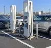 Two white electric vehicle charging stations are shown in a parking lot with cars parked nearby. Náhled obrázku produktu