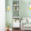 A tall, narrow white bookshelf with wooden legs is placed next to a light gray sofa in a room with a pale green wall. A potted cactus sits on a wooden stool to the left of the bookshelf. Produktbild-Vorschau