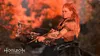 A woman with red hair is aiming a bow and arrow in a natural setting with an orange sky. Produktbild-Vorschau
