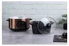A rose gold slow cooker with a black insert and glass lid sits on a table in front of a white brick wall. Náhled obrázku produktu