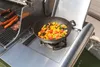 A wok pan filled with chopped vegetables is placed on a side burner of a gas grill, next to sausages and a piece of meat on the main grill grates. Podgląd zdjęcia produktu