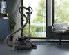 A vacuum cleaner is shown in a modern living room with large windows overlooking a garden. Produktbild-Vorschau