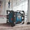 A construction site with a Bosch radio on a wooden surface and a worker on a ladder in the background. Produktbild-Vorschau