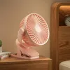 A pink clip-on fan sits on a wooden nightstand next to a book and a small potted plant. Produktbild-Vorschau