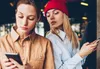 Two young women are looking at their phones, with one woman looking over the other's shoulder. Anteprima dell’immagine prodotto